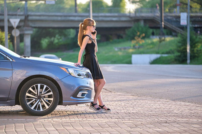Young mom standing by car on phone, captured outside near a trampoline park in a sunny urban setting.