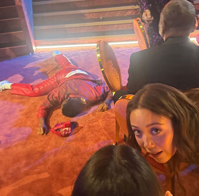 Celebrity in a red suit lying on the floor with awkward reactions during the 2026 Grammy Awards event.