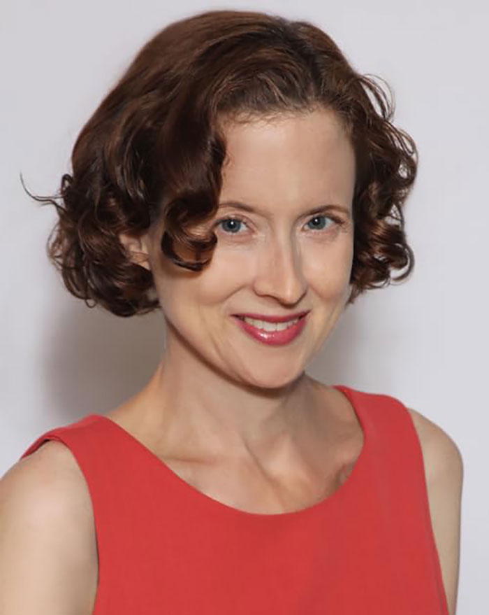 Actress from Grey's Anatomy smiling, wearing a red dress with short curly brown hair against a plain background.