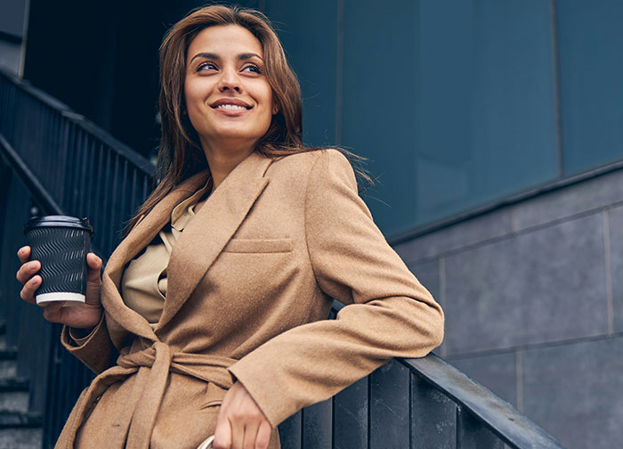 Confident divorce lawyer in camel coat holding coffee cup outdoors near modern office building staircase.