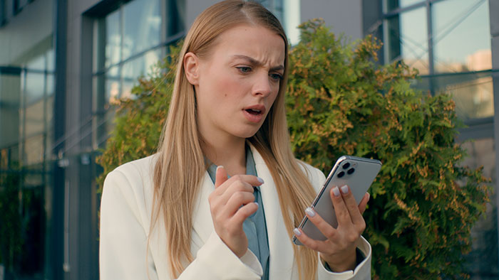 Woman at airport looking upset while checking phone, reacting to missing carry-on and friend ditching her.