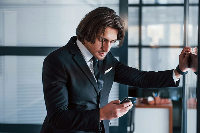 Man in black suit looking at phone, appearing upset after wife reveals plans for brother's wedding outfit.