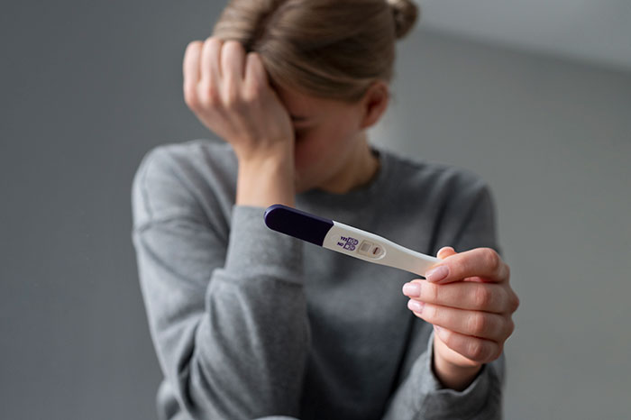 Woman in gray sweatshirt showing positive pregnancy test, expressing confusion and concern about partner being pregnant.