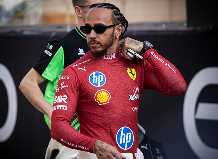Lewis Hamilton in a Ferrari racing suit and sunglasses showing confident body language at a motorsport event.