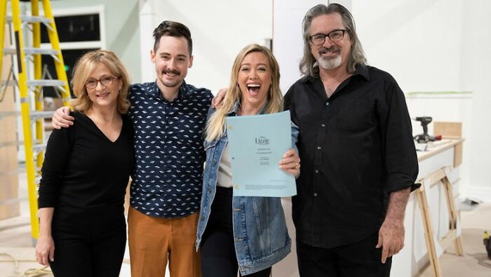 Four cast members smiling on a set holding a Lizzie McGuire script amid fan fury over Disney&rsquo;s controversial decision.