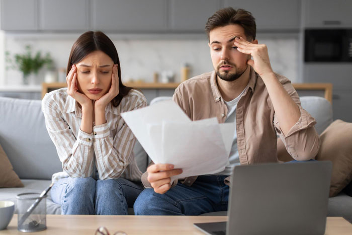 Couple looking stressed on a couch, man holding documents and woman feeling humiliated about locked money box situation.