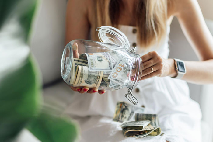 Woman holding a glass money box with hundred dollar bills inside, symbolizing money control and trust issues.