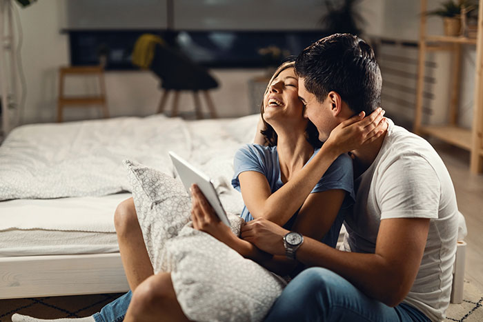 Couple with long hair sharing a joyful moment on bed as the husband looks suspicious about shower time.
