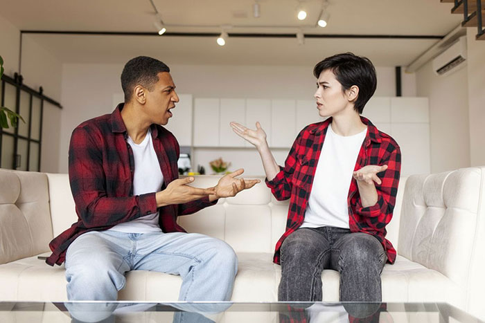 Couple having a heated argument on a couch, suspicious husband questioning his long-haired partner indoors.