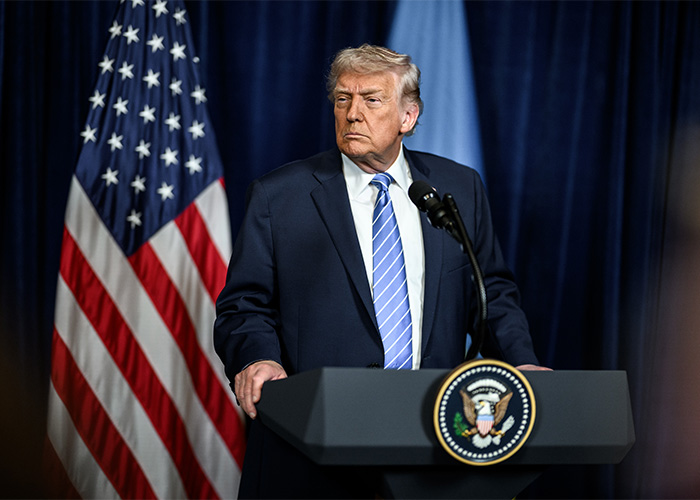 Former President Trump at a podium with the US flag, related to MAGA influencer detained by ICE after deportation remarks.