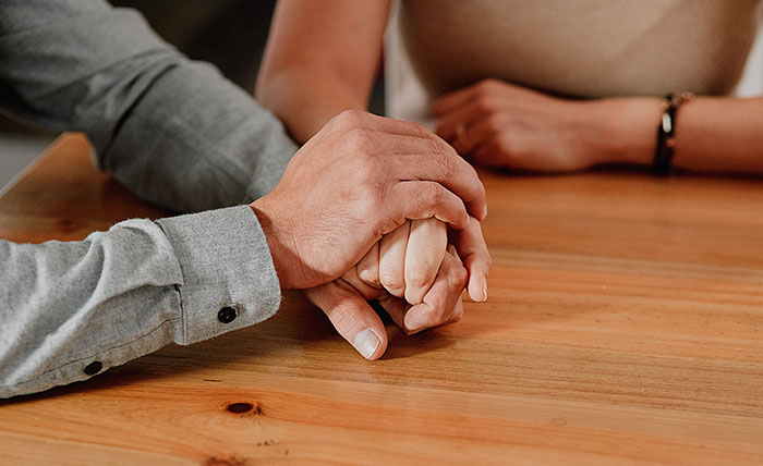 A husband gently holding his wife's hand, struggling to accept her weight gain while sitting at a wooden table.