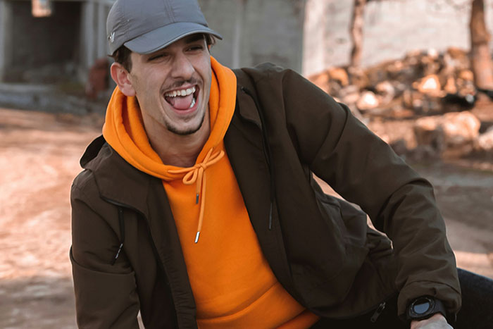 Young groom in orange hoodie and gray cap laughing while pranking brother-in-law’s girlfriend at wedding event outdoors.