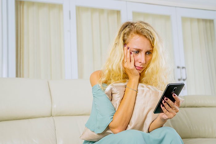 Woman looking depressed while sitting on a couch, holding a pillow and checking her phone in a man poly relationship context