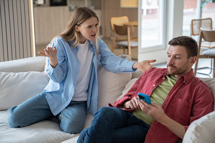 Woman volunteering boyfriend for free labor looks upset while man refuses and stays focused on his phone on the couch