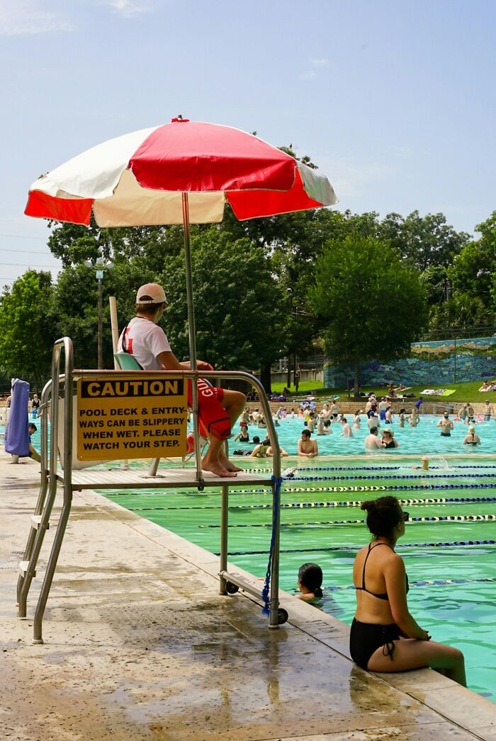 Lifeguard under red umbrella watches crowded pool where people relax and swim on a sunny day with trees in background.