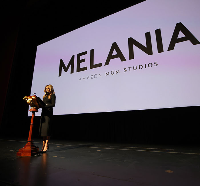 Speaker standing at podium on stage in front of large screen displaying Melania Amazon MGM Studios logo during theater event.