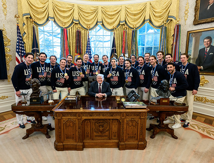 US women's hockey players wearing medals posing with former president in the Oval Office after Olympic victory celebration