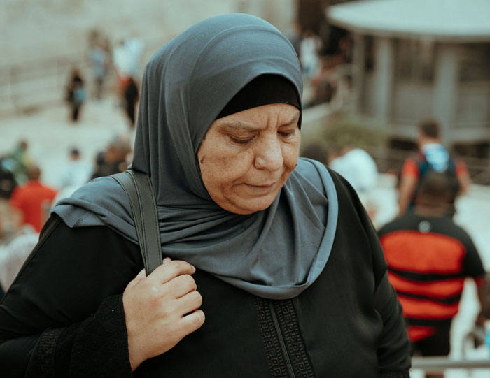 Muslim mother-in-law in a gray hijab looking down with a concerned expression in a busy outdoor setting. Muslim mother-in-law in a gray hijab looking down with a concerned expression in a busy outdoor setting.