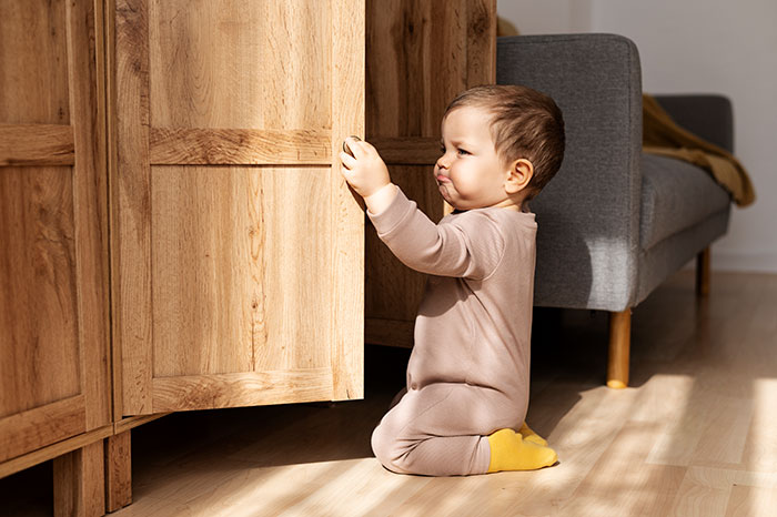 Toddler reaching for lower cabinet door in a living room, highlighting the need to keep meds out of kid's reach for safety.