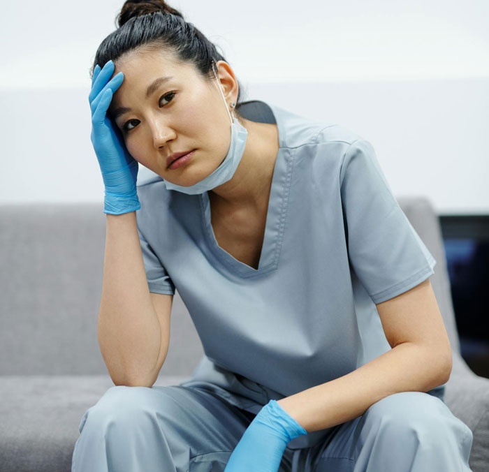 Stressed nurse in scrubs and gloves sitting on a couch, holding her head, symbolizing HIPAA-related work stress.