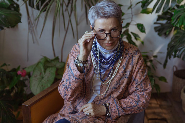 Older woman adjusting glasses sitting indoors surrounded by plants, reflecting a nurse's stress about HIPAA privacy.