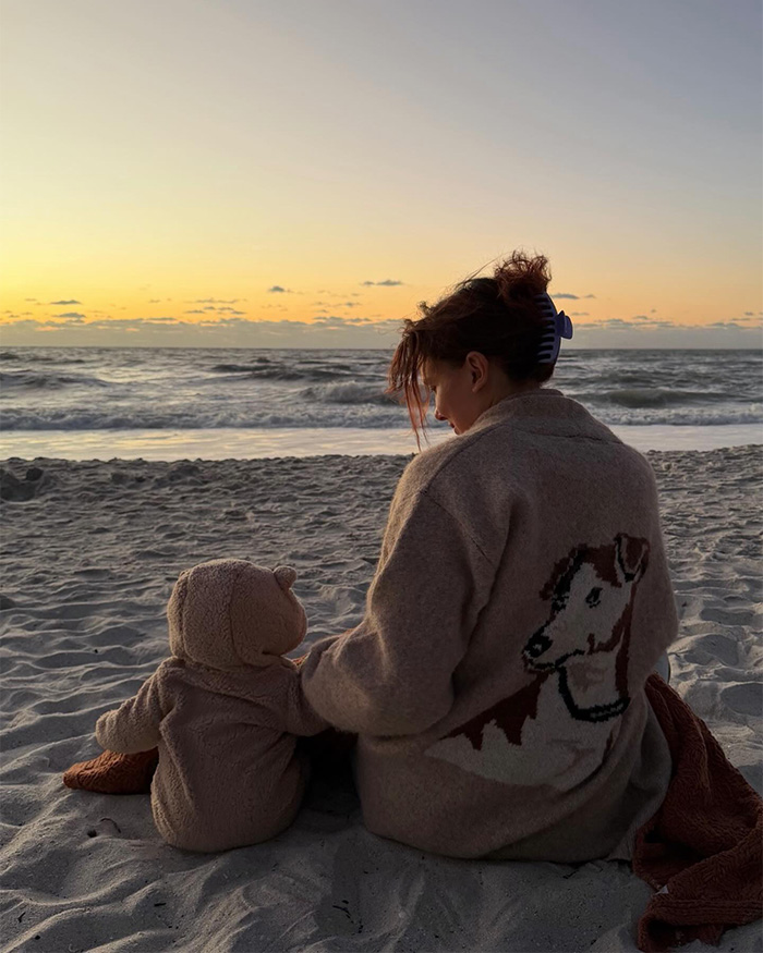 Millie Bobby Brown sitting on the beach at sunset with her baby daughter dressed in matching cozy outfits.