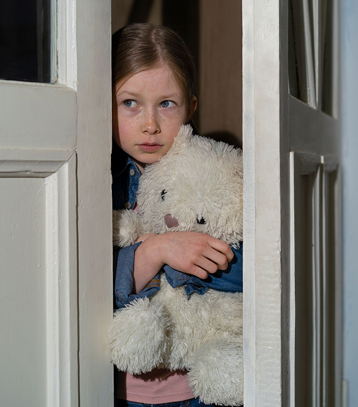 Young girl clutching a teddy bear nervously peeking through a door, illustrating disturbing student confessions to teachers.