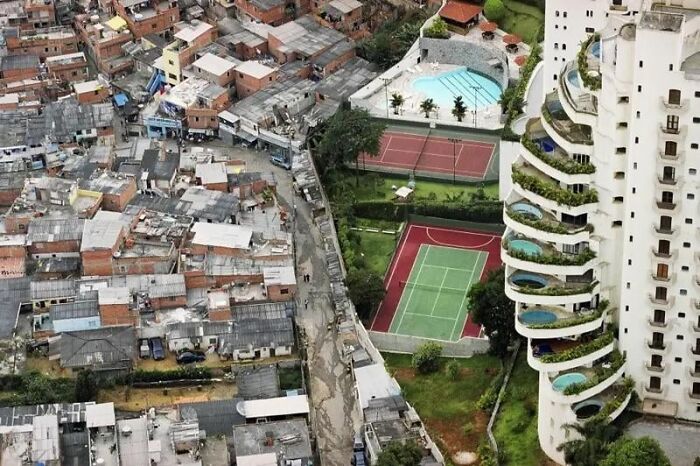 A stark contrast between a crowded slum and luxurious apartments with pools and sports courts, dystopian nightmare scene.