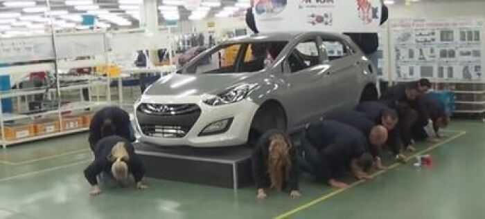 Workers crawling on floor inside factory resembling a dystopian nightmare scene with a white car on display.
