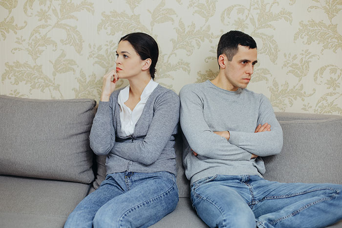 Wife and lazy hubby sitting apart on couch, wife upset and husband avoiding chores after inviting mom to help.
