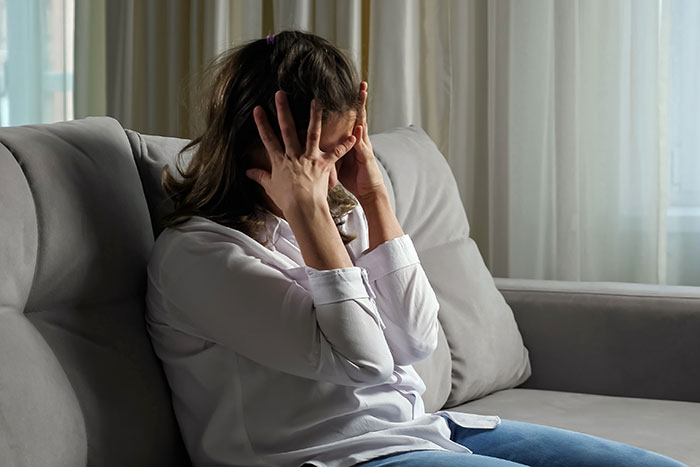 Woman sitting on couch in distress, covering her face, illustrating postpartum boundaries retaliation emotion.