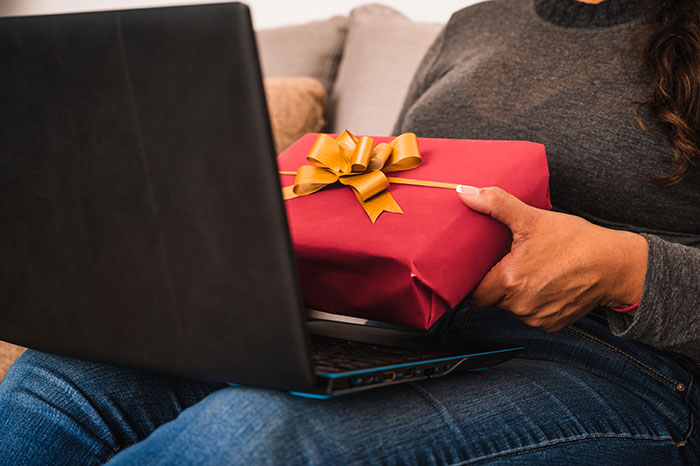 Person sitting with a laptop on their lap, holding a red gift box with a golden ribbon, highlighting postpartum boundaries.