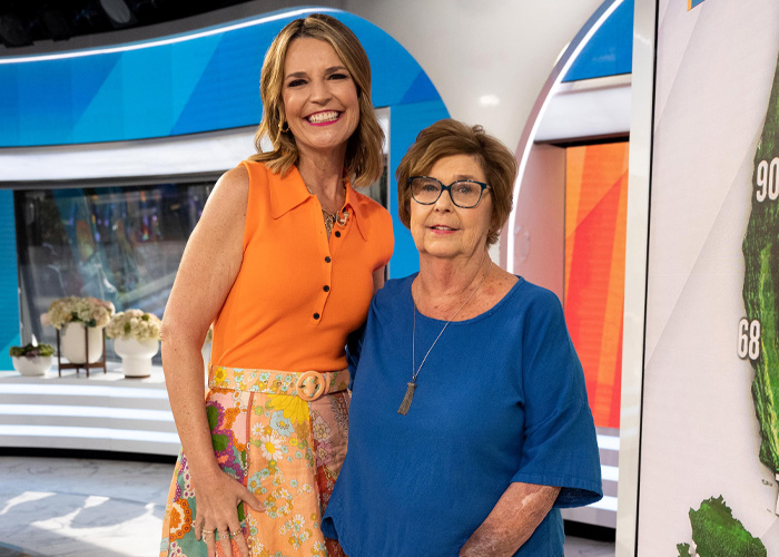 Two women, one in an orange top and floral skirt, and the other in blue, standing together in a brightly lit studio setting.