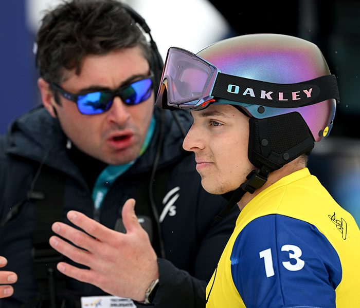 Athlete wearing iridescent Oakley helmet and yellow bib number 13 listening to coach during 2026 Winter Olympics event.