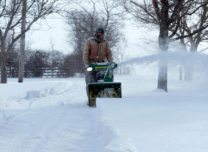 Man using a snowblower clearing snow on a driveway surrounded by trees on a snowy winter day.