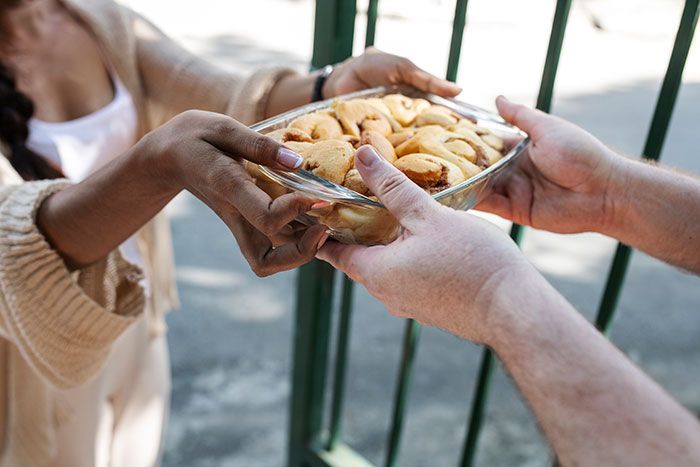 Hands exchanging a glass dish of cookies outside, symbolizing stress and paranoia with an unhinged neighbor nearby.