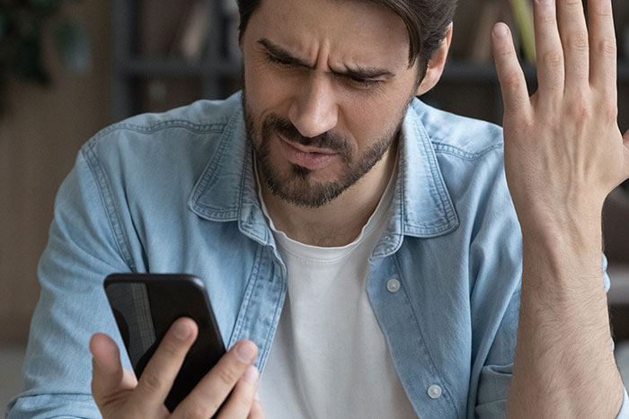 Man stressed and paranoid looking at phone, frustrated by unhinged neighbor causing anxiety at home.