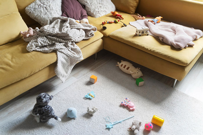 Living room with messy play area, toys scattered on rug, and blankets on yellow sectional sofa after playdate chaos.
