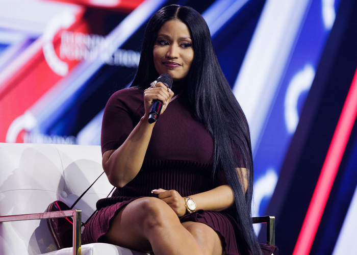 Nicki Minaj sitting on stage holding a microphone with long black hair and a dark purple dress during an event.