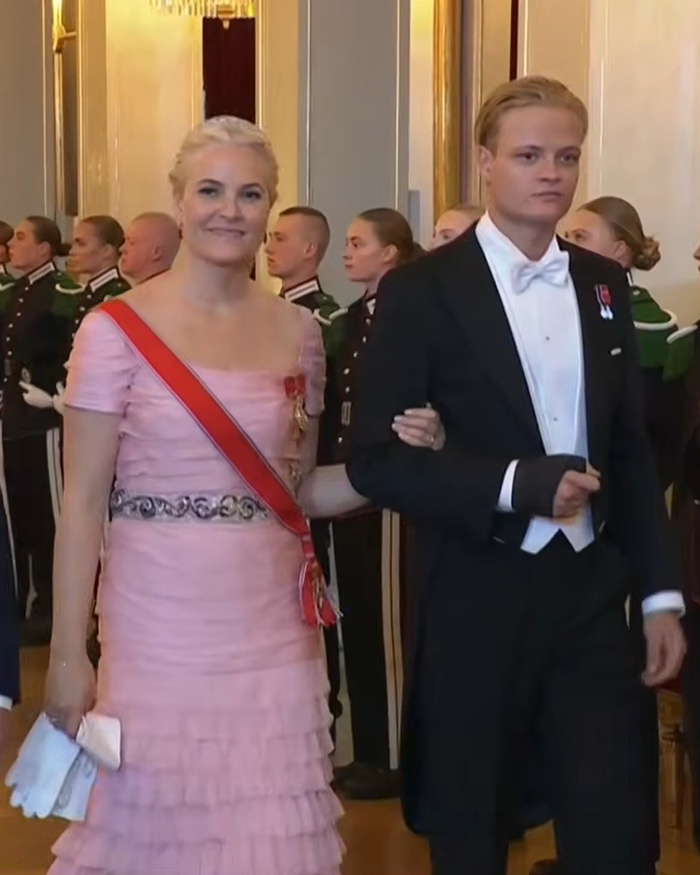 Son of Norway's Crown Princess in formal attire, arm-in-arm with a woman in a pink dress at a ceremonial event.