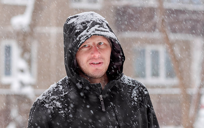 Man wearing a black hooded jacket covered in snow standing outside during a snowstorm with buildings in the background.