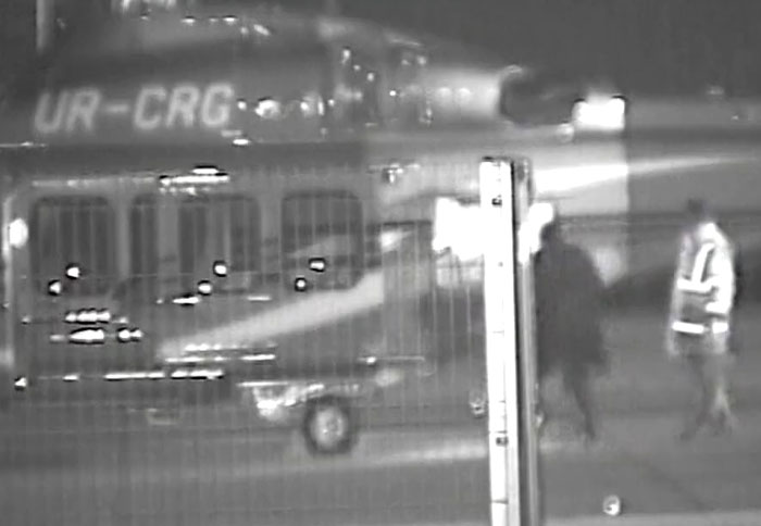 Black and white powerful photo capturing people walking near a lighted helicopter at night in an outdoor setting.