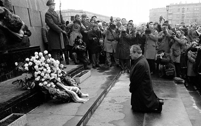 Man kneeling before a memorial wreath while photographers and guards observe in a powerful photo from around the world.