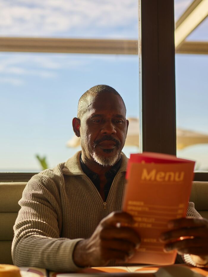 Middle-aged man reading a menu in a sunlit cafe, reflecting on respect in relationships and partner dynamics.
