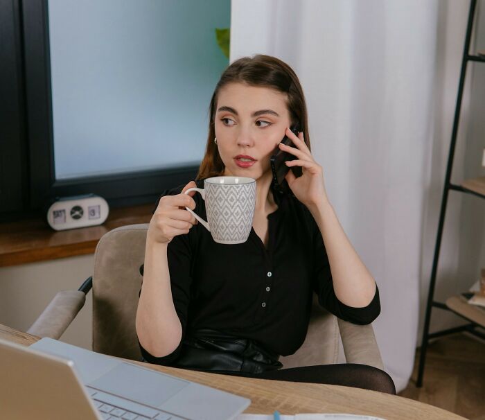 Young woman at desk holding coffee mug and talking on phone, showing a toxic workplace moment with stressed expression.