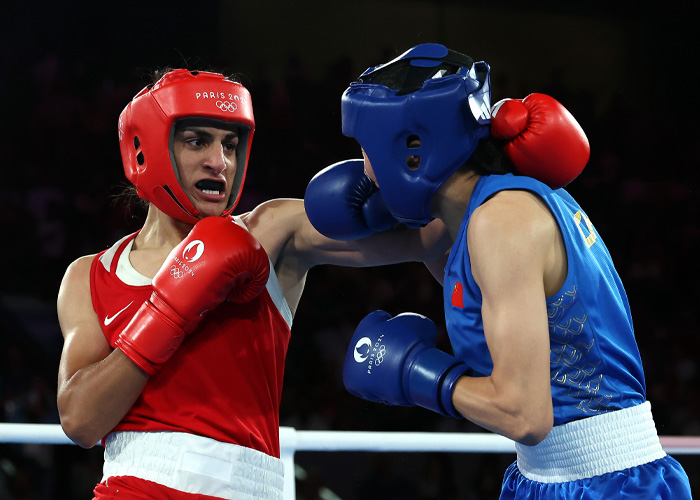 Female boxer wearing red headgear and gloves landing a punch on an opponent in blue during a competitive boxing match.