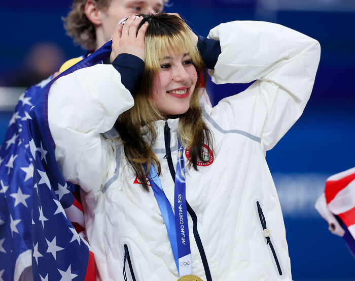 US figure skater smiling with a gold medal and American flag draped, celebrating during award ceremony.