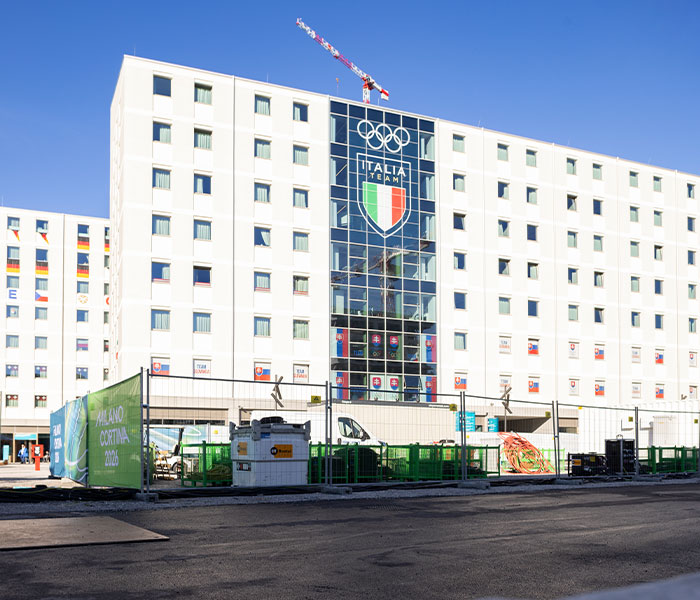 Olympic athlete housing under construction at the 2026 Winter Olympics with Italy team emblem on building facade.