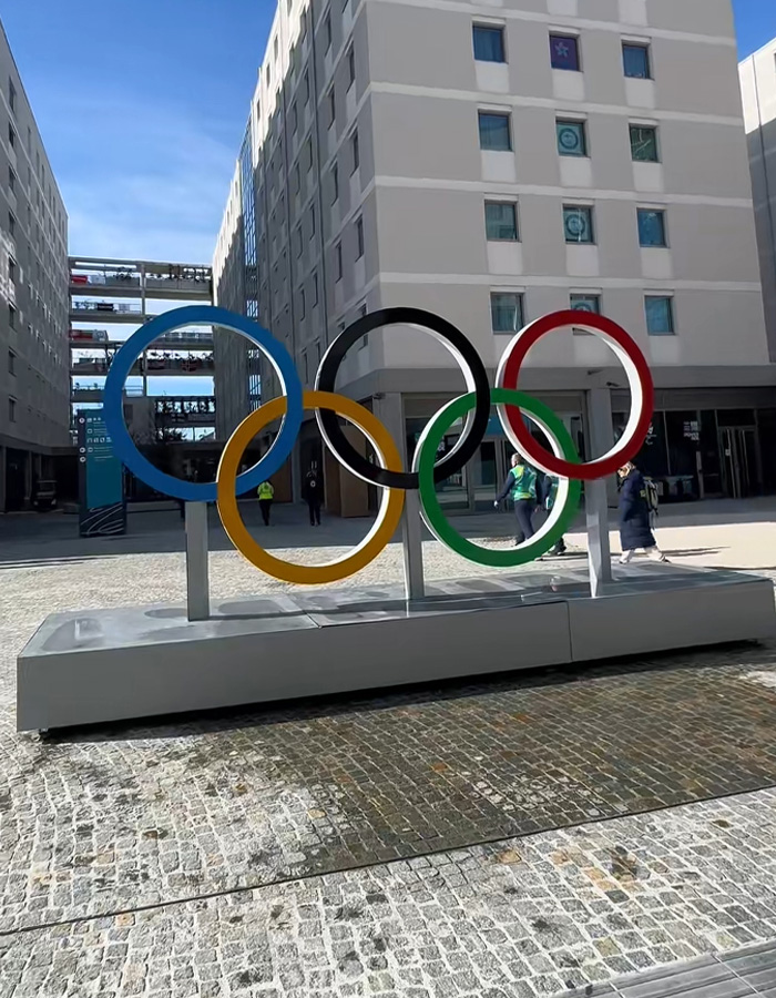 Olympic Village courtyard with large colorful Olympic rings sculpture and athletes walking near modern buildings on a sunny day