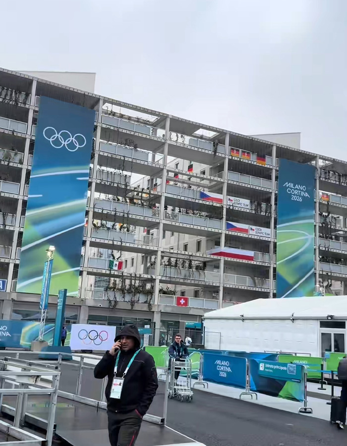 Olympic Village exterior with banners and flags, showing athletes and staff during a cold day at the Winter Games event.